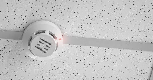 A small, white smoke detector placed on a ceiling. The ceiling is a drop ceiling and is colored a light gray.